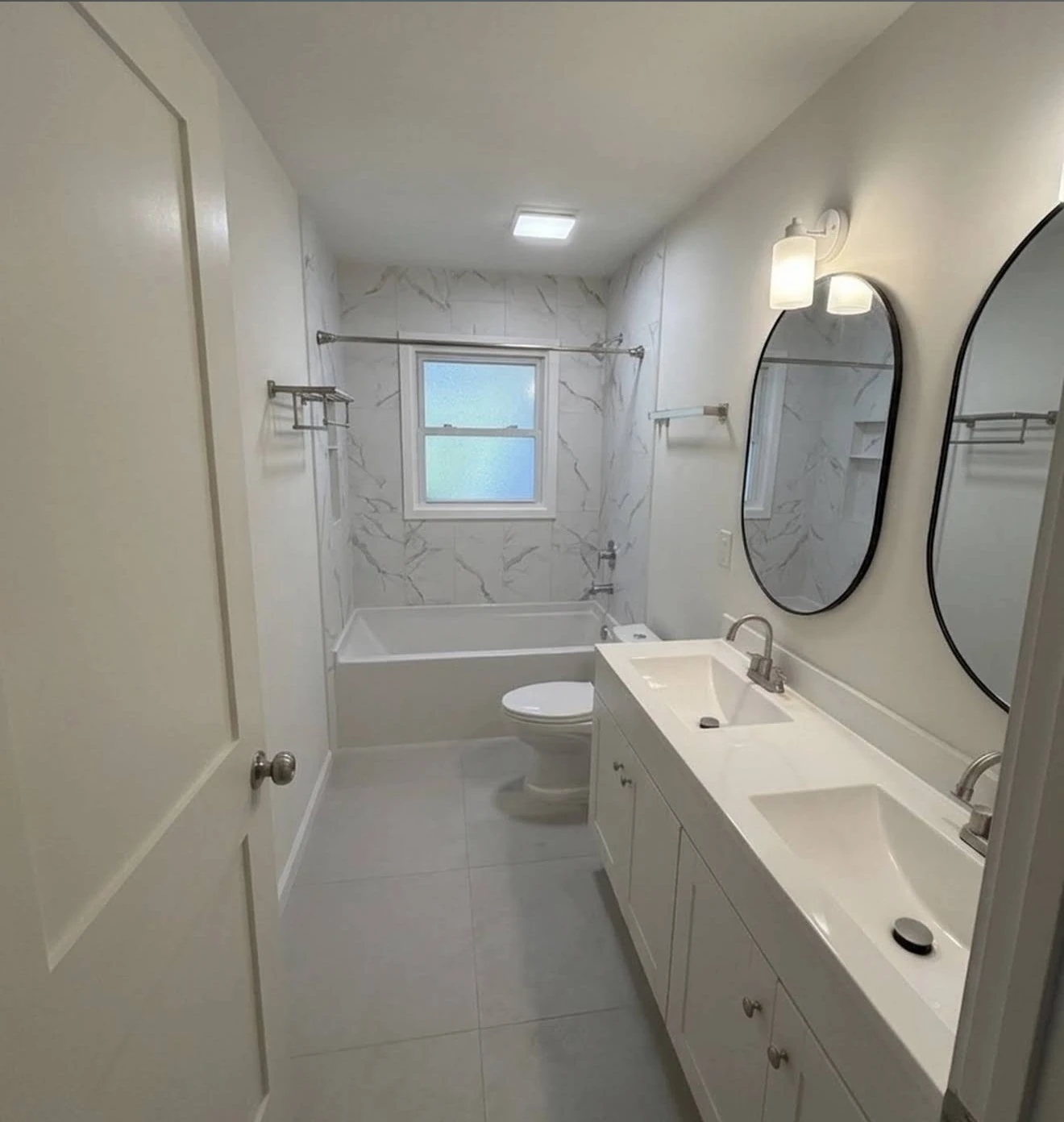 Renovated bathroom with a double vanity, two oval mirrors and a white marble-look tub surround